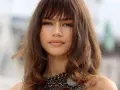 Primo piano di una donna con un **paffuto taglio capelli viso tondo**. Capelli castani mossi con frangia, labbra lucide e una collana a catena spessa.