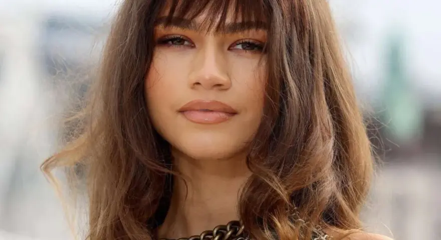 Primo piano di una donna con un **paffuto taglio capelli viso tondo**. Capelli castani mossi con frangia, labbra lucide e una collana a catena spessa.