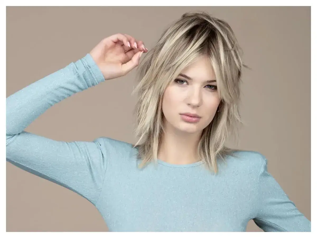 Modella con un taglio scalato medio corto biondo, indossa un maglione azzurro scintillante.