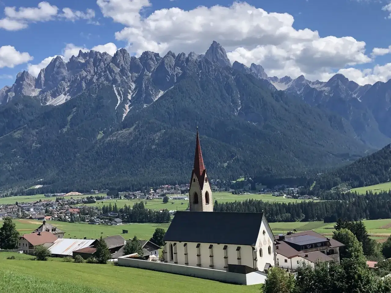 Chiesa con campanile rosso in primo piano, sullo sfondo le montagne maestose e i paesi pi&ugrave; belli della Val Pusteria.