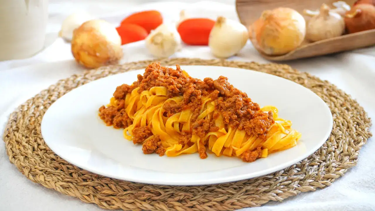 Un piatto invitante di tagliatelle al rag&ugrave;, con ingredienti freschi come pomodori e cipolle sullo sfondo.