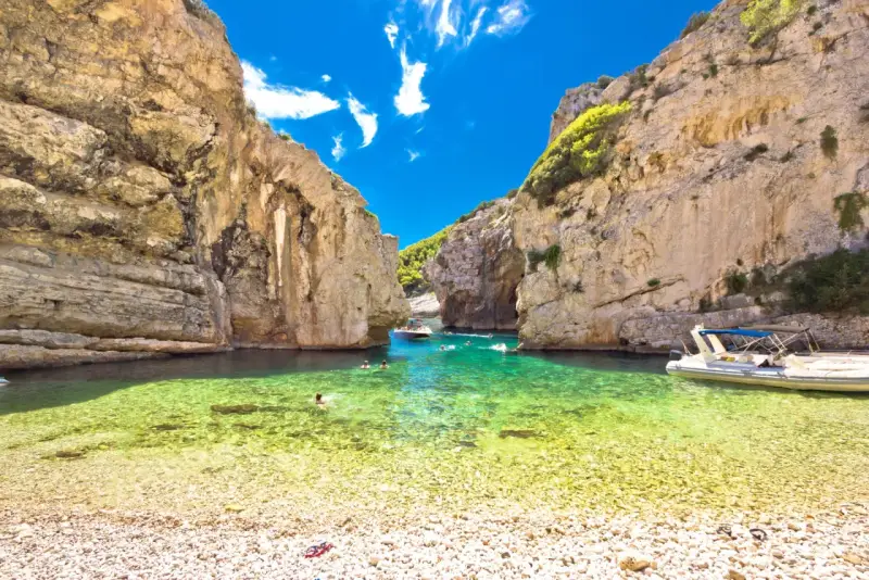 Spiaggia di ciottoli e acque cristalline tra le rocce dell'isola di Lissa, con persone che nuotano e barche.