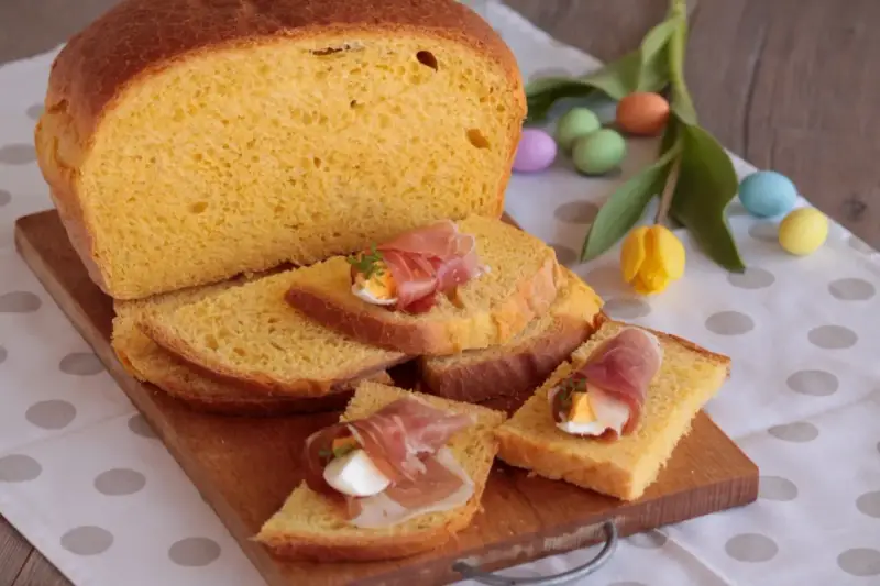 Fetta di torta al formaggio umbra ricetta originale, guarnita con uovo sodo e prosciutto crudo, su tagliere di legno, con uova colorate e tulipano.